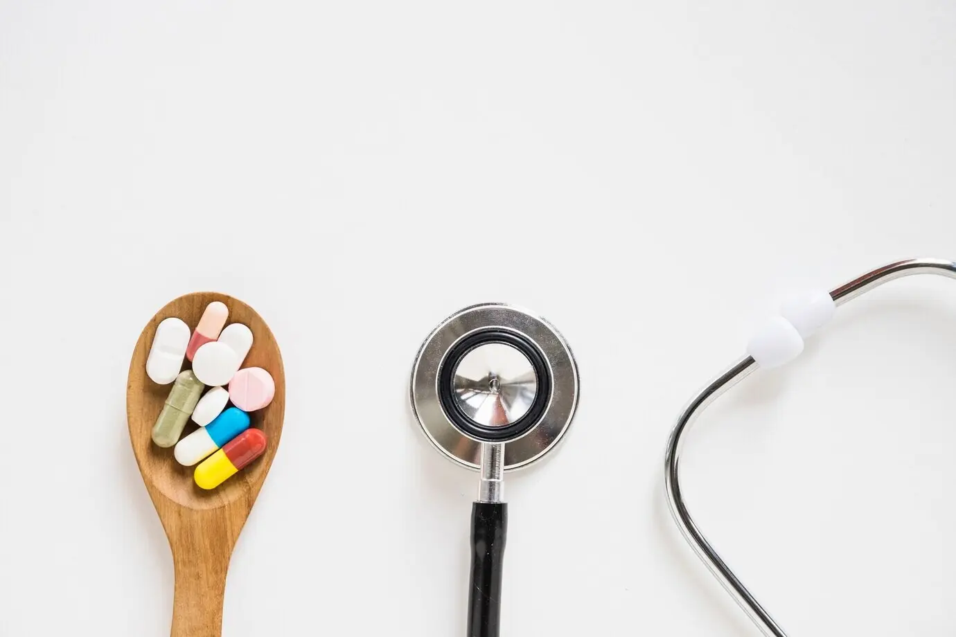 Auf weißem Hintergrund sind ein Holzlöffel mit bunten Pillen und ein Stethoskop zu sehen.