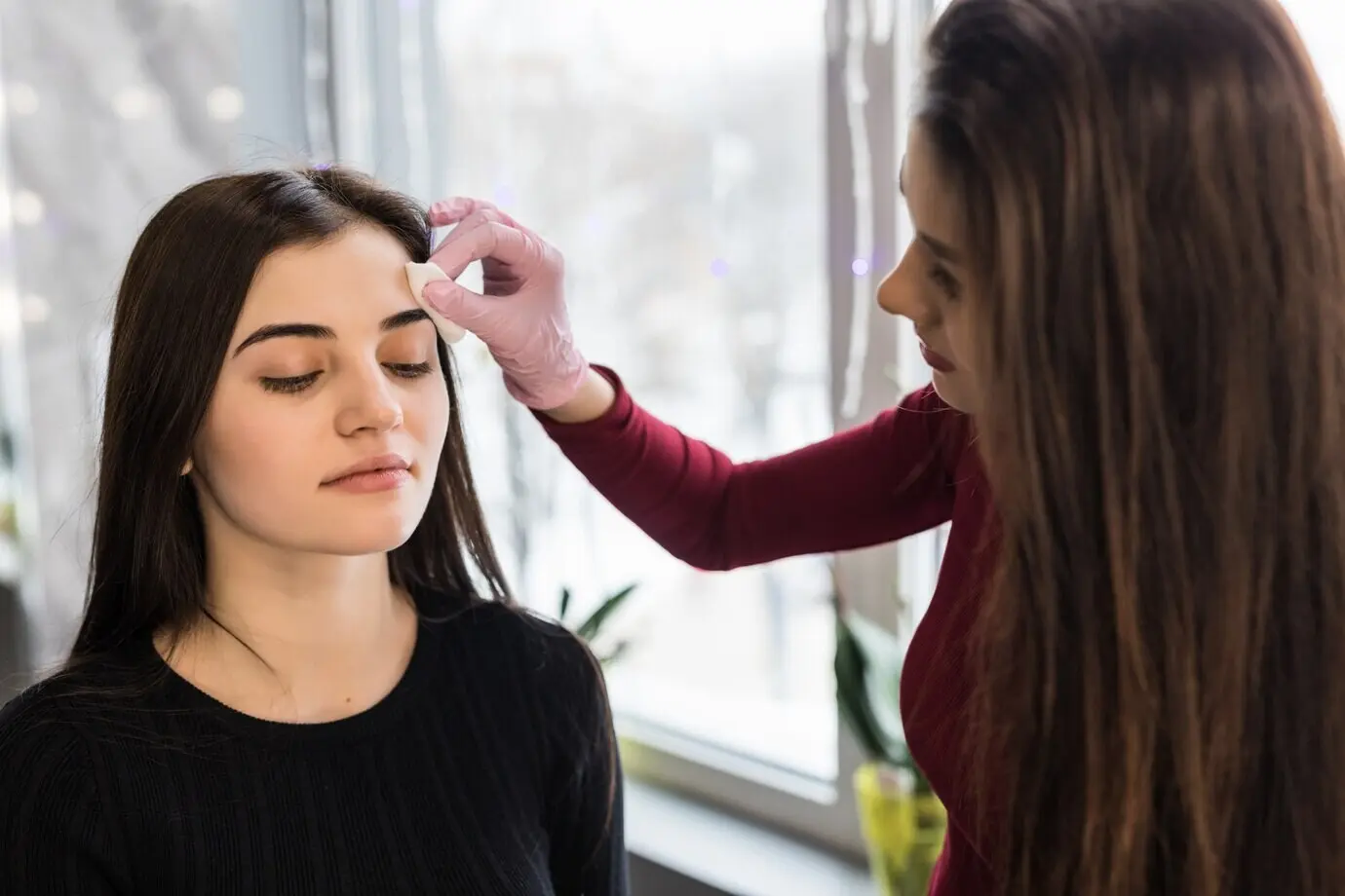 Gekonnt trägt ein/e Visagist/in bei einer jungen Frau mit dunklen Haaren Augenbrauen-Make-up auf.